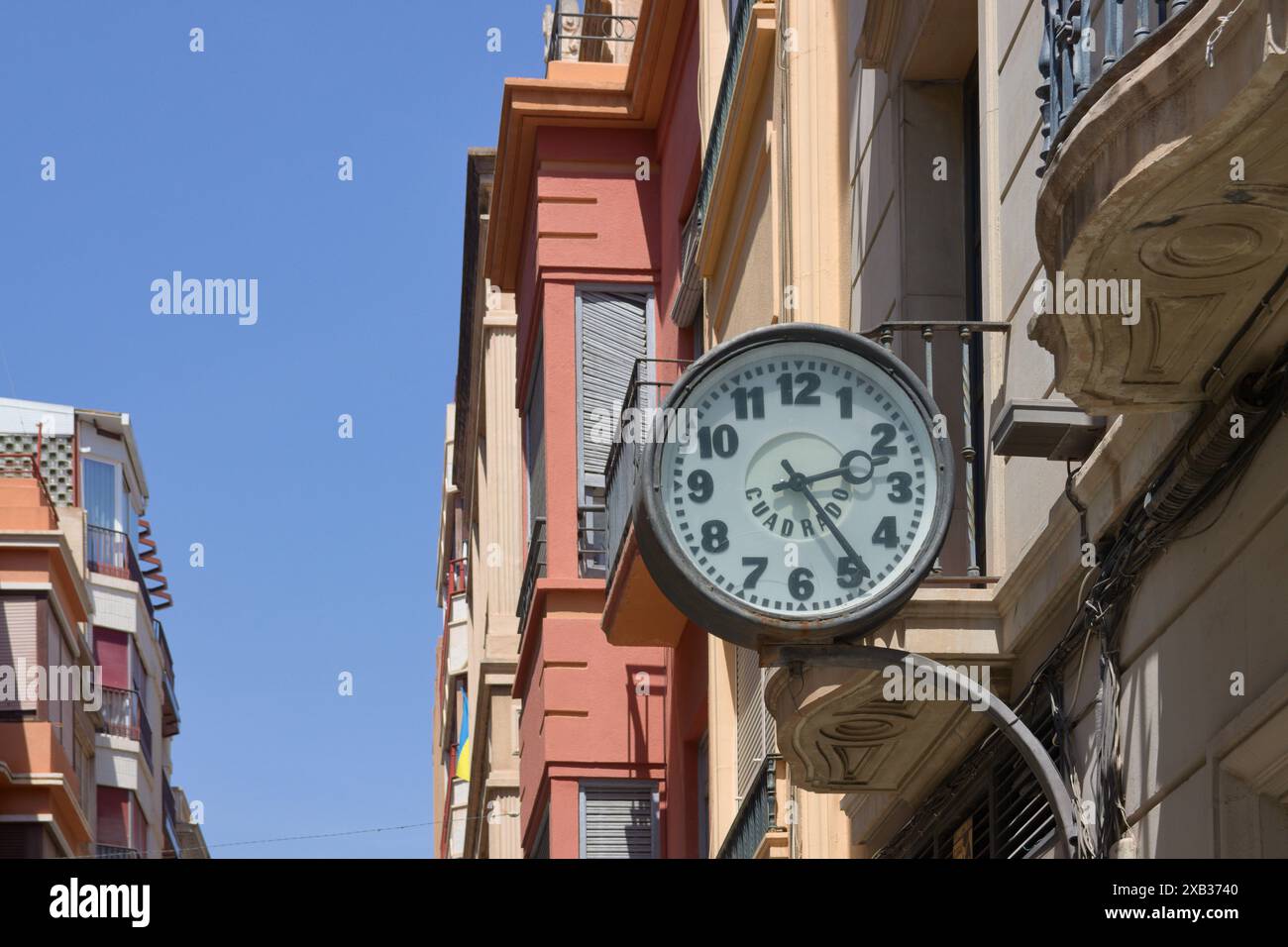 Primo piano di un vecchio orologio analogico che mostra le 14:00, appeso a uno degli edifici di una strada Foto Stock Primo piano di un vecchio orologio analogico che mostra le 14:00, appeso a uno degli edifici di una strada Foto Stock