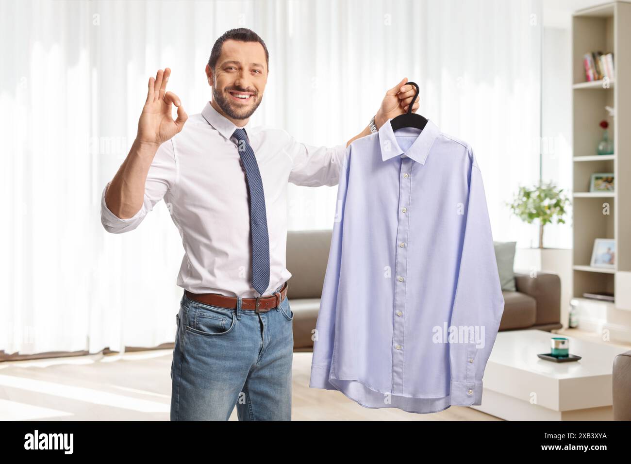 Uomo d'affari che tiene una camicia su un gancio a casa in un soggiorno e che gesta ok Foto Stock