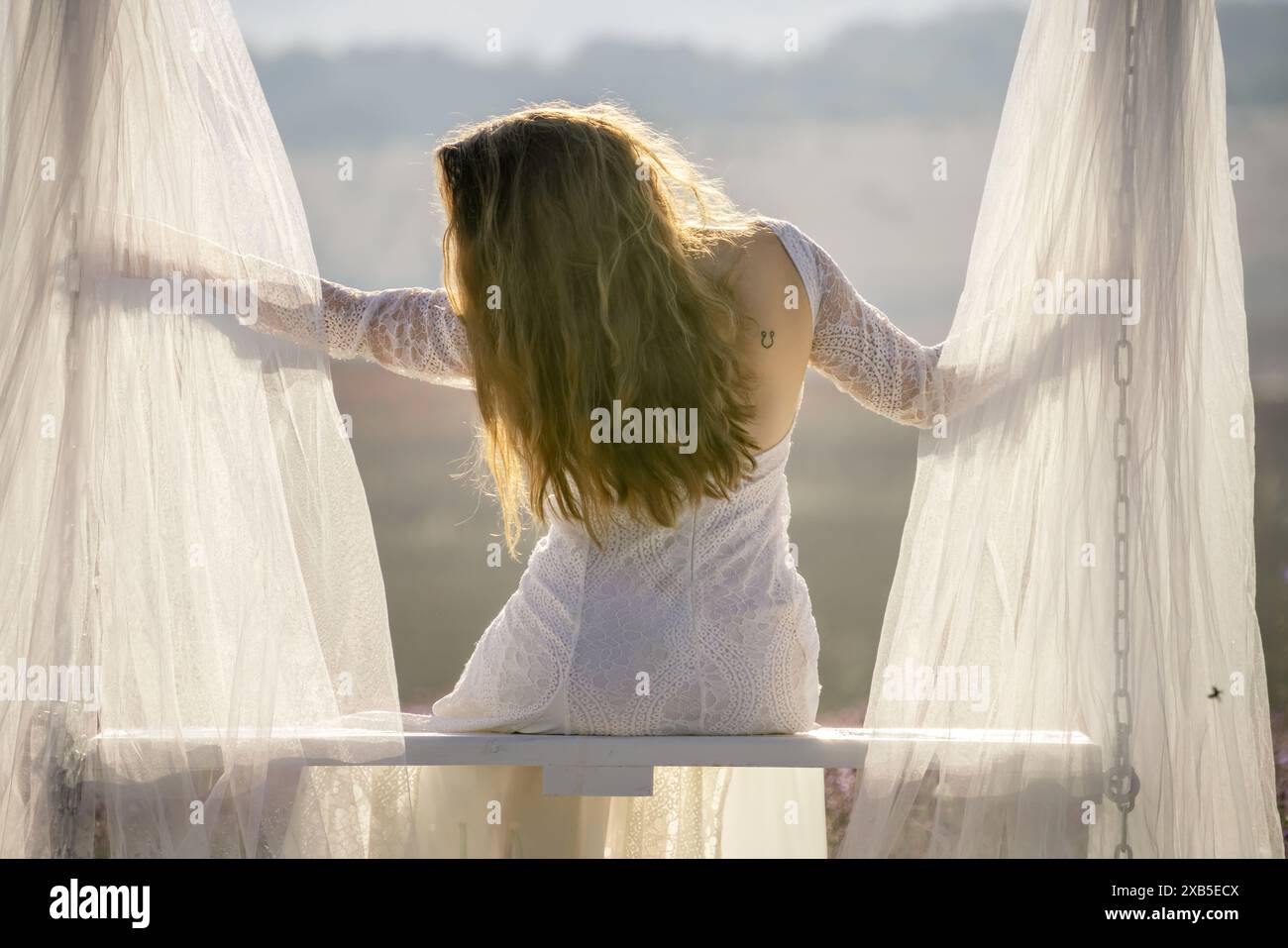 Una donna con un abito bianco siede su una panchina con una tenda bianca dietro di lei. Il sipario è aperto, rivelando una splendida vista sulle montagne. La donna Foto Stock