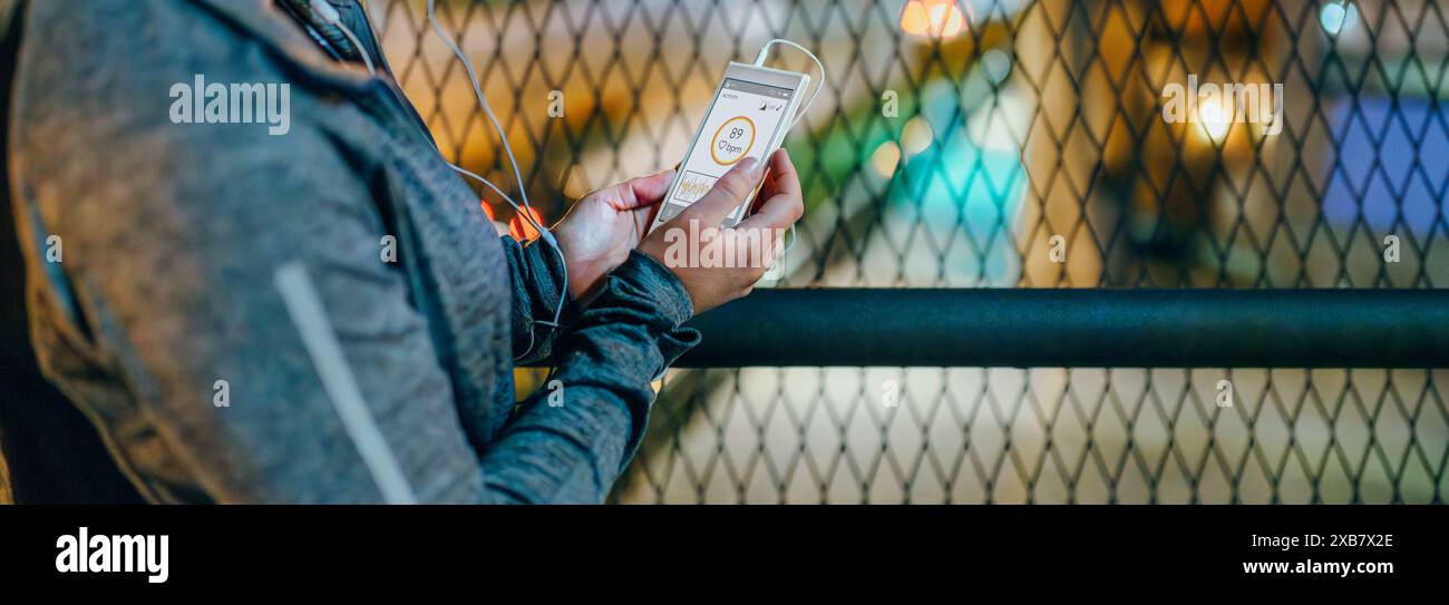 Striscione di donna runner che tiene in mano un telefono cellulare con cardiofrequenzimetro sullo schermo dopo l'allenamento in città di notte. Atleta femminile irriconoscibile che tiene c Foto Stock