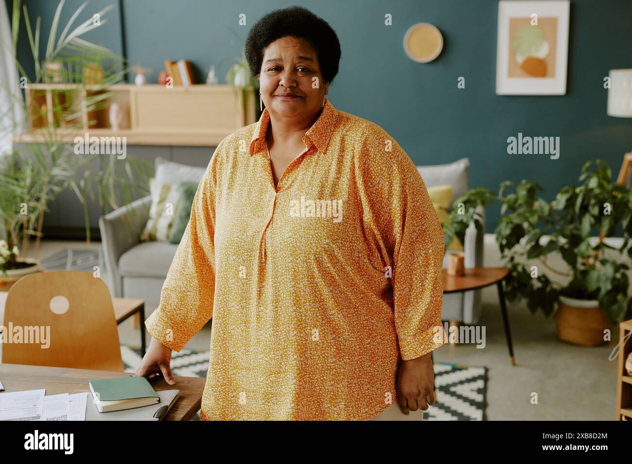 Donna d'affari afroamericana matura in piedi accanto alla scrivania mentre lavorava in remoto e sorrideva con un sorriso stanco Foto Stock