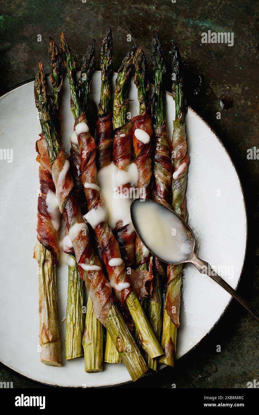 Asparagi confezionati con pancetta serviti su un piatto bianco con un coltello e una forchetta vintage, accompagnati da una piccola ciotola di salsa. La pancetta è croccante, e. Foto Stock