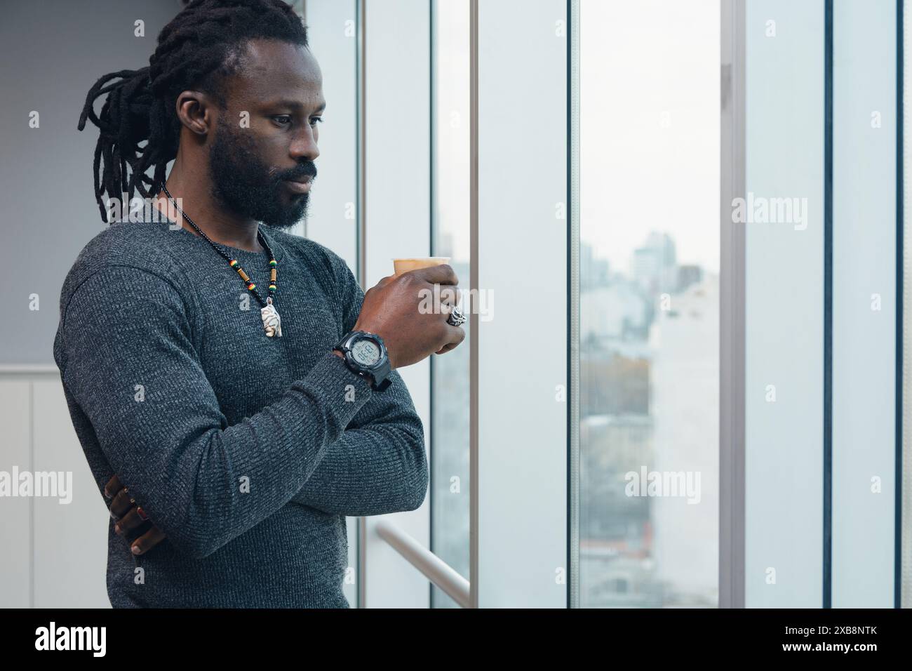 Adulto lavoratore d'ufficio africano uomo è in piedi vicino alla finestra pensando alle strategie aziendali. L'uomo indossa un maglione, un orologio e una collana è lo Foto Stock
