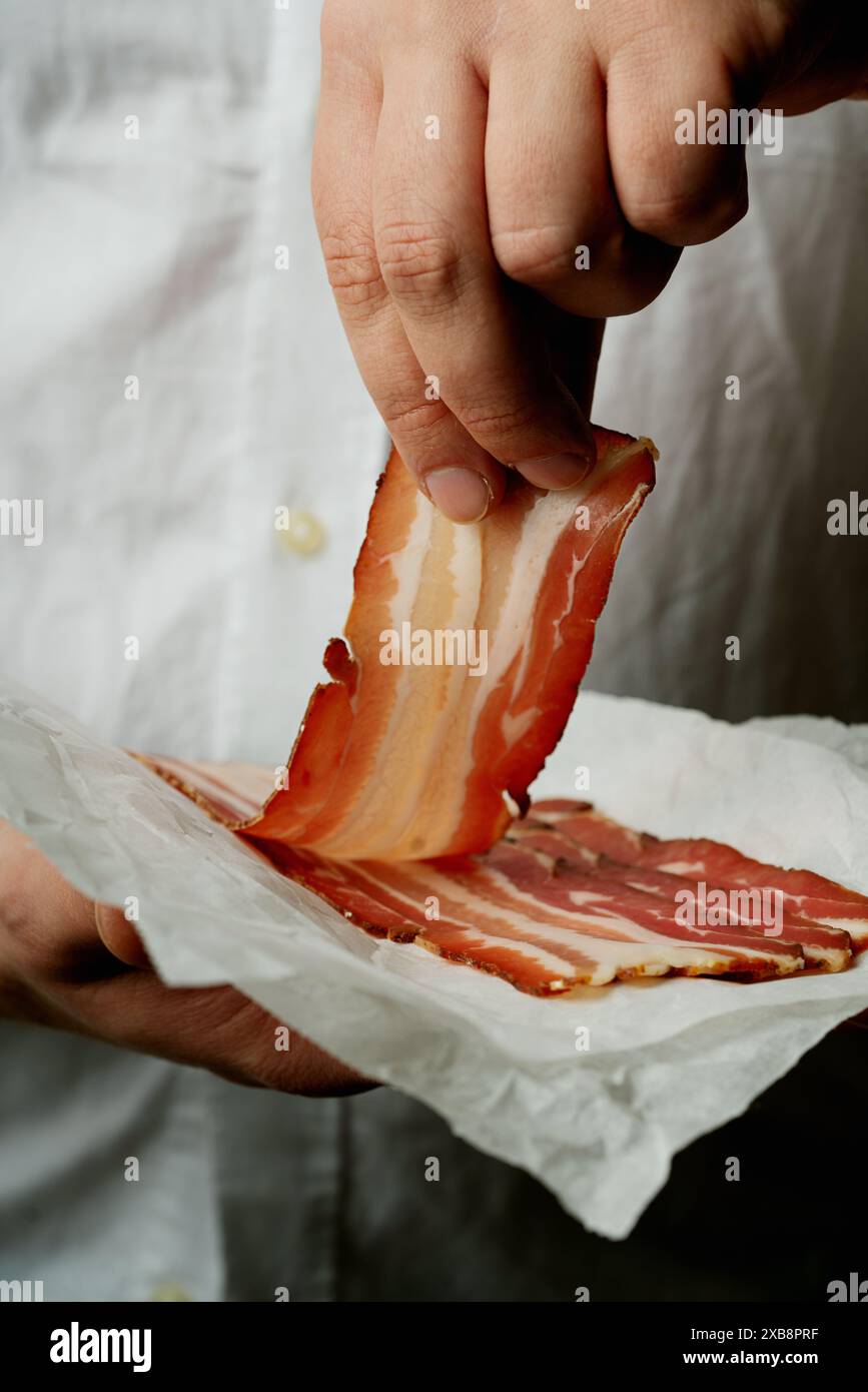 Primo piano di una mano che tiene una fetta sottile di pancetta marmorizzata, sollevata da una pila di fette su carta pergamena. La scena si concentra sulla trama e sui dettagli Foto Stock