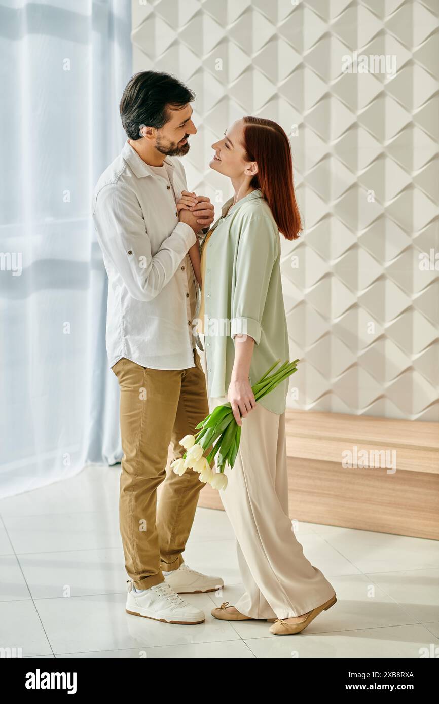 Una donna rossa e un uomo barbuto abbracciano in una stanza adornata da fiori colorati, godendosi momenti di qualità insieme a casa. Foto Stock