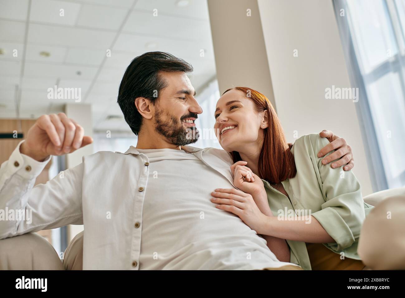 Una bella coppia adulta, una donna rossa e un uomo barbuto, si siedono insieme su un divano moderno nel loro appartamento, godendosi del tempo di qualità. Foto Stock