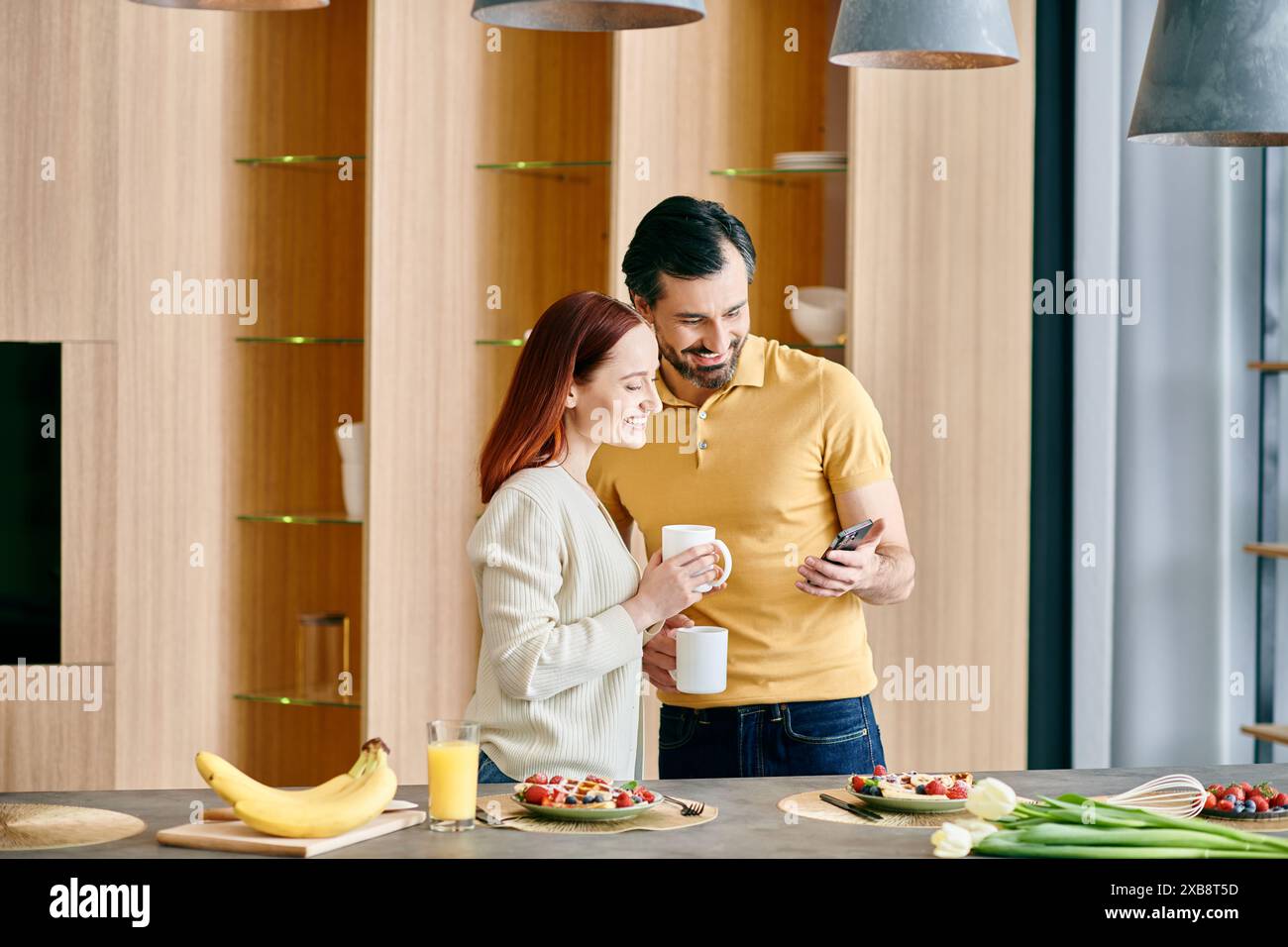 Una donna rossa e un uomo barbuto guardano il loro telefono, in piedi in una cucina moderna, godendo del tempo di qualità a casa. Foto Stock