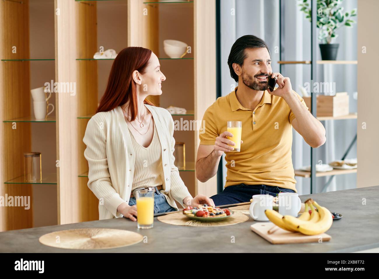 Donna rossa e uomo barbuto che chiacchierano al telefono mentre facevano colazione in un appartamento moderno. Foto Stock