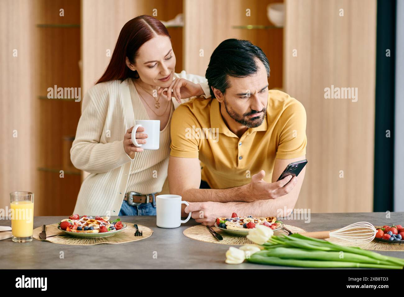 Una donna rossa e un uomo barbuto si sono invischiati nei loro telefoni mentre condividono la colazione in un appartamento moderno. Foto Stock