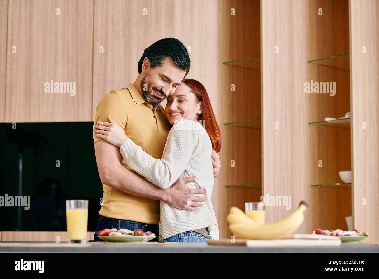 Una donna rossa e un uomo barbuto condividono un caldo abbraccio in una cucina moderna, ridendo e adorando il momento insieme. Foto Stock
