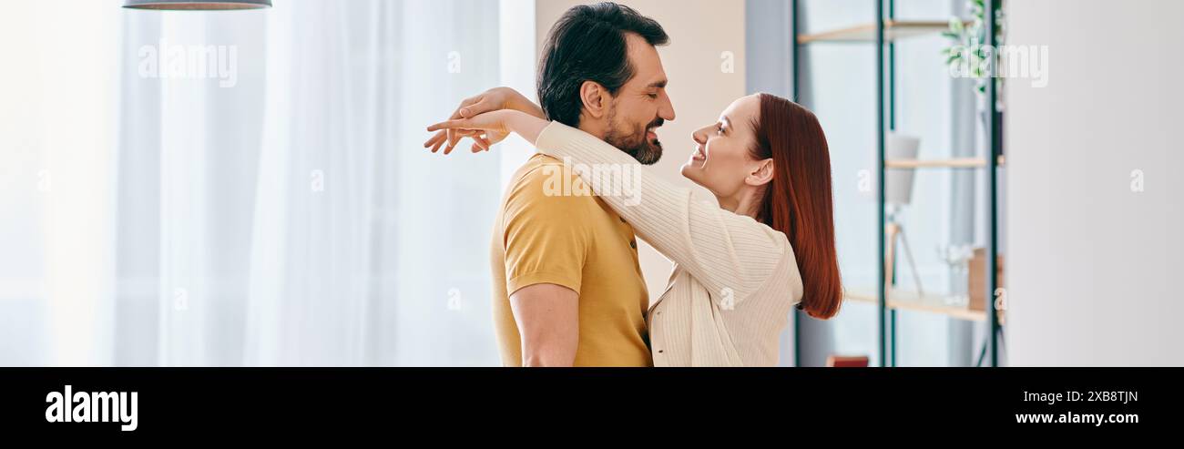 Una donna rossa e un uomo barbuto condividono un caldo abbraccio in un accogliente soggiorno, creando un bellissimo momento di intimità e connessione. Foto Stock