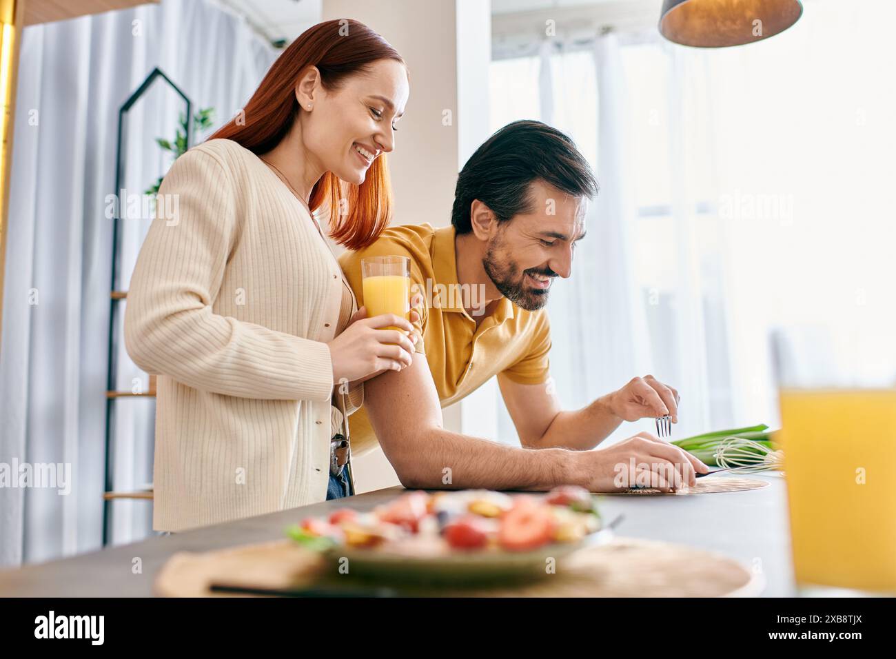 Una donna rossa e un uomo barbuto cucinano un pasto insieme in una cucina moderna, condividendo un momento speciale preparando il cibo. Foto Stock