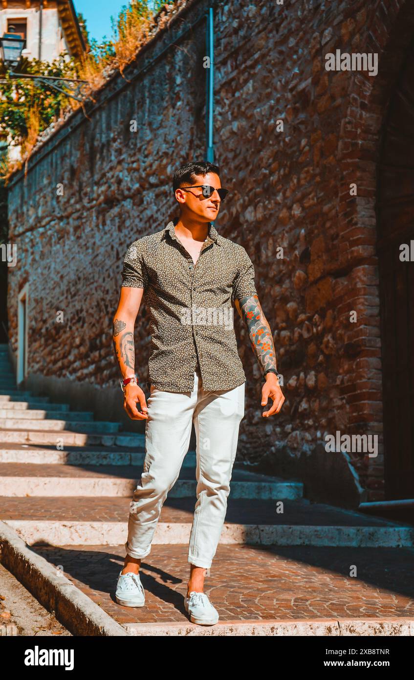 Giovane uomo elegante con occhiali da sole e camicia in abito che passeggiano in un ambiente urbano Foto Stock