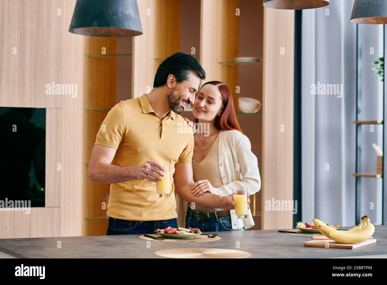 Una donna rossa e un uomo barbuto condividono un momento accogliente, in piedi insieme in una cucina moderna. Foto Stock