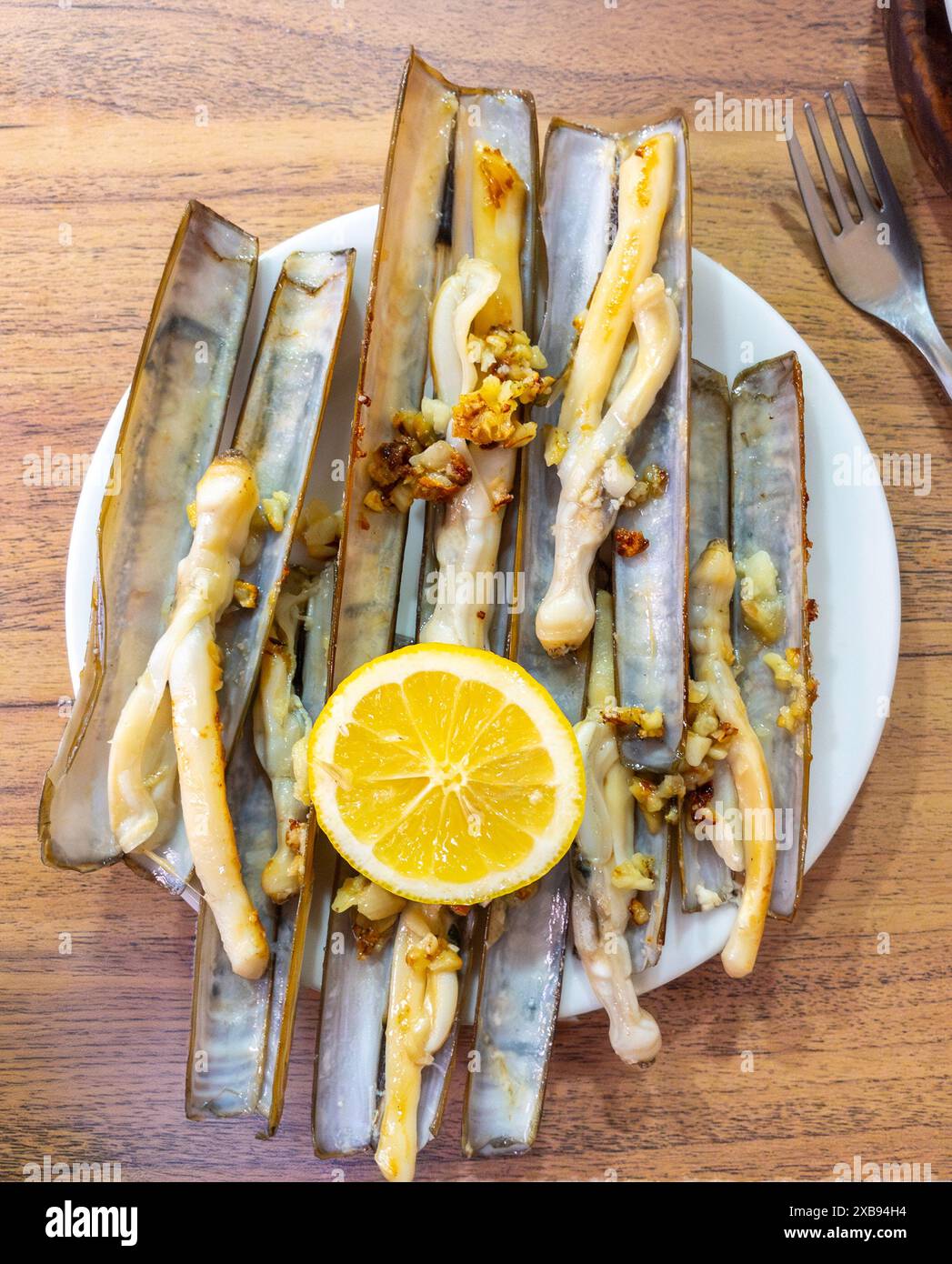 Vongole grigliate in stile spagnolo, guarnite con aglio e una fetta di limone, servite su un tavolo di legno. Foto Stock