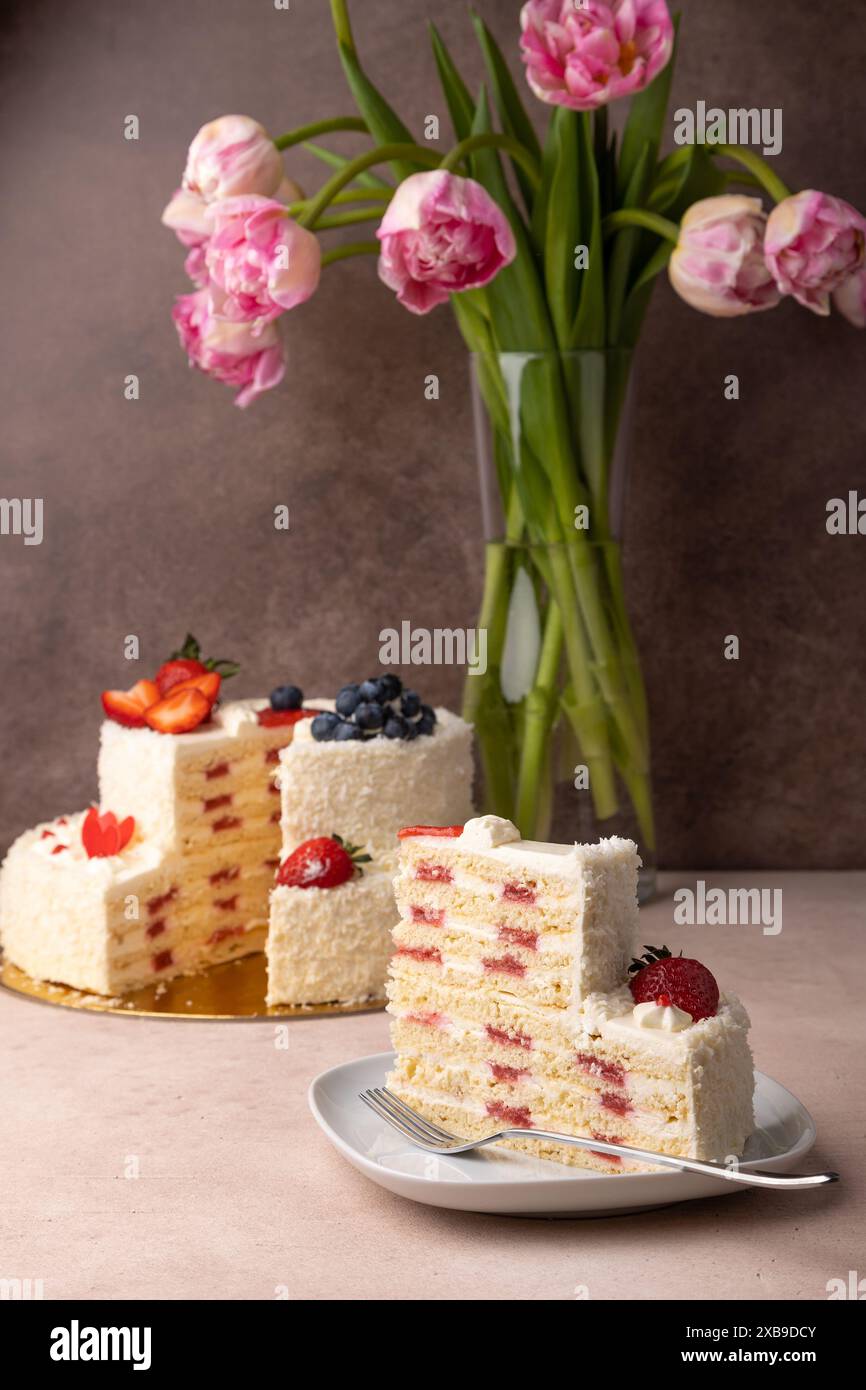 Torta di compleanno alla fragola e al cocco con panna e frutti di bosco freschi. Una fetta di torta di compleanno a strati fatta in casa. Sullo sfondo c'è un bouquet di rosa t Foto Stock