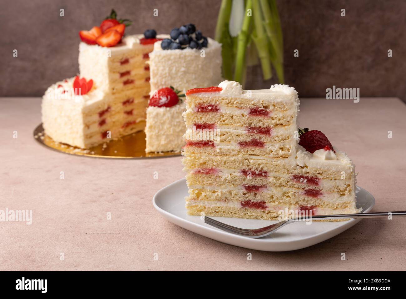 Torta di compleanno alla fragola e al cocco con panna e frutti di bosco freschi. Una fetta di torta di compleanno a strati fatta in casa. Sullo sfondo c'è un bouquet. Foto Stock