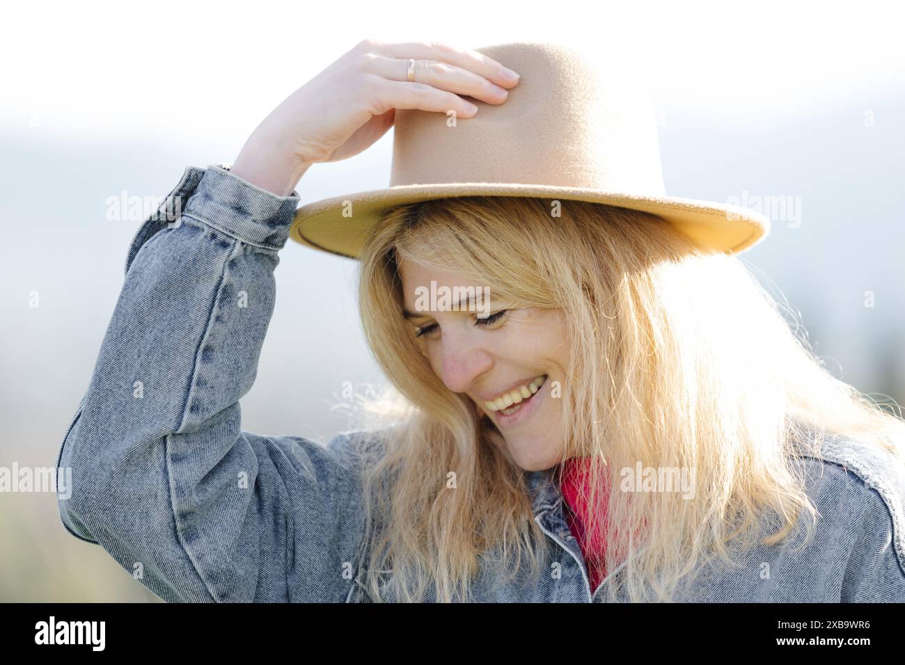 Una donna bionda che indossa un cappello e una giacca in denim sorride mentre guarda in basso in una giornata di sole. Foto Stock