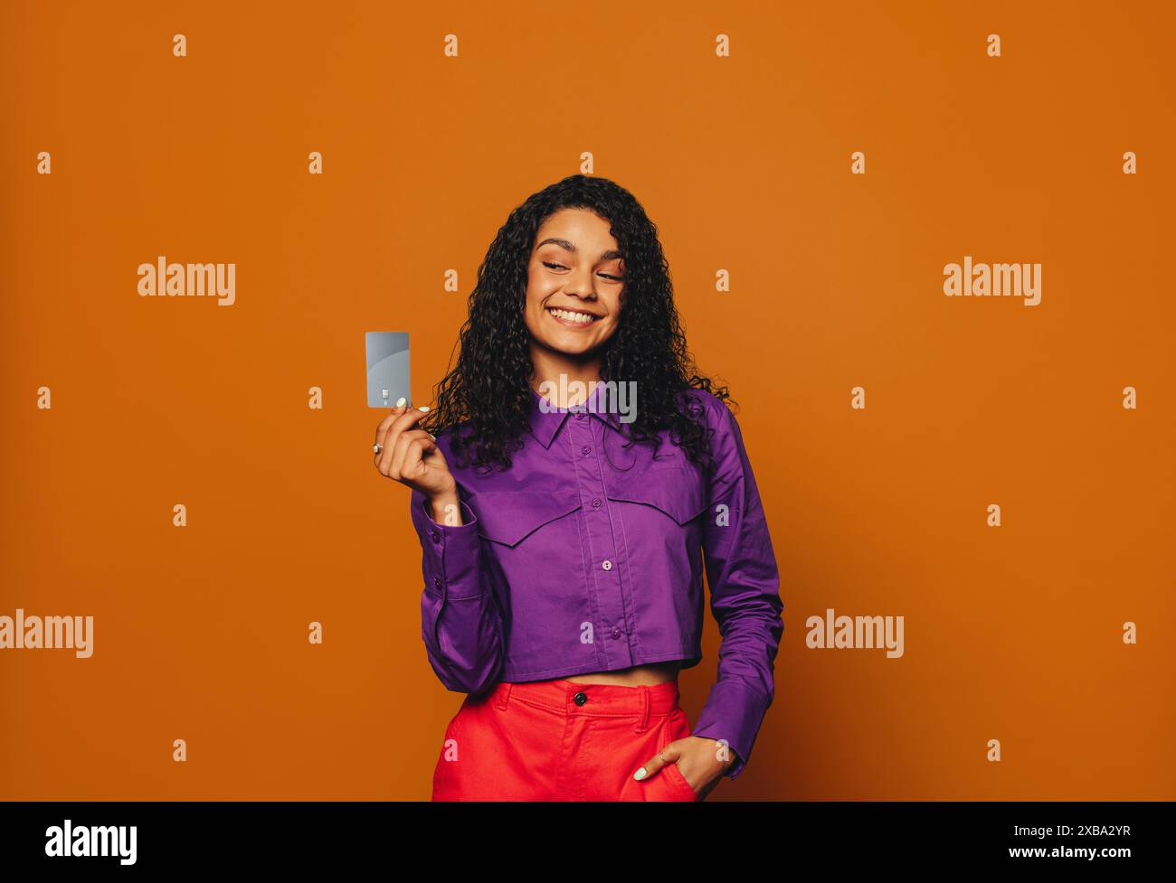 Donna dalla moda allegra si staglia su un vivace sfondo arancione. Ha in mano una carta di credito, sorridendo mentre paga senza contatto. Foto Stock