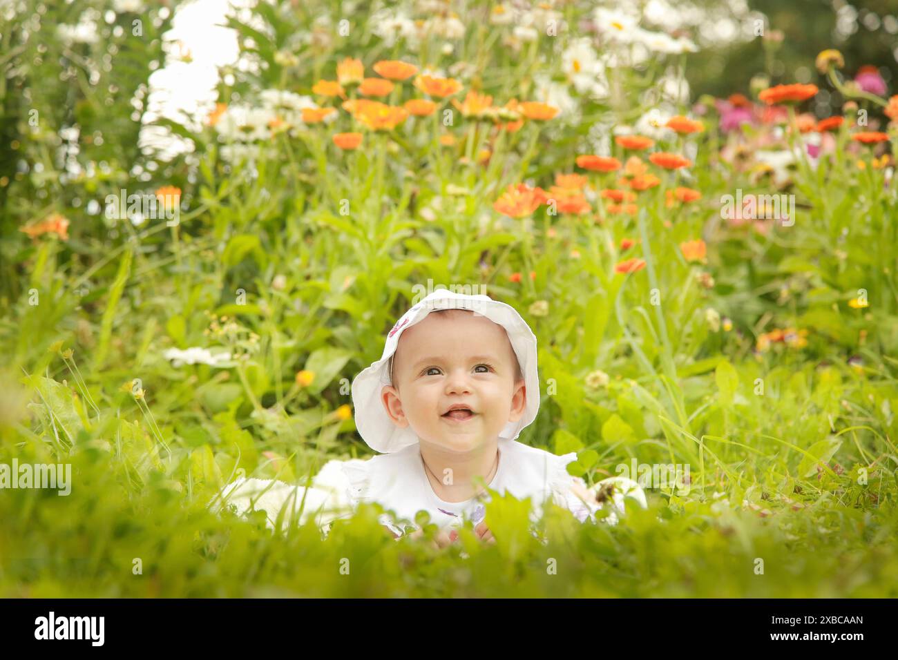 Un bambino sorridente che indossa un cappello bianco si siede in un giardino pieno di fiori colorati e vegetazione lussureggiante in una giornata di sole, la Bielorussia. Minsk Foto Stock