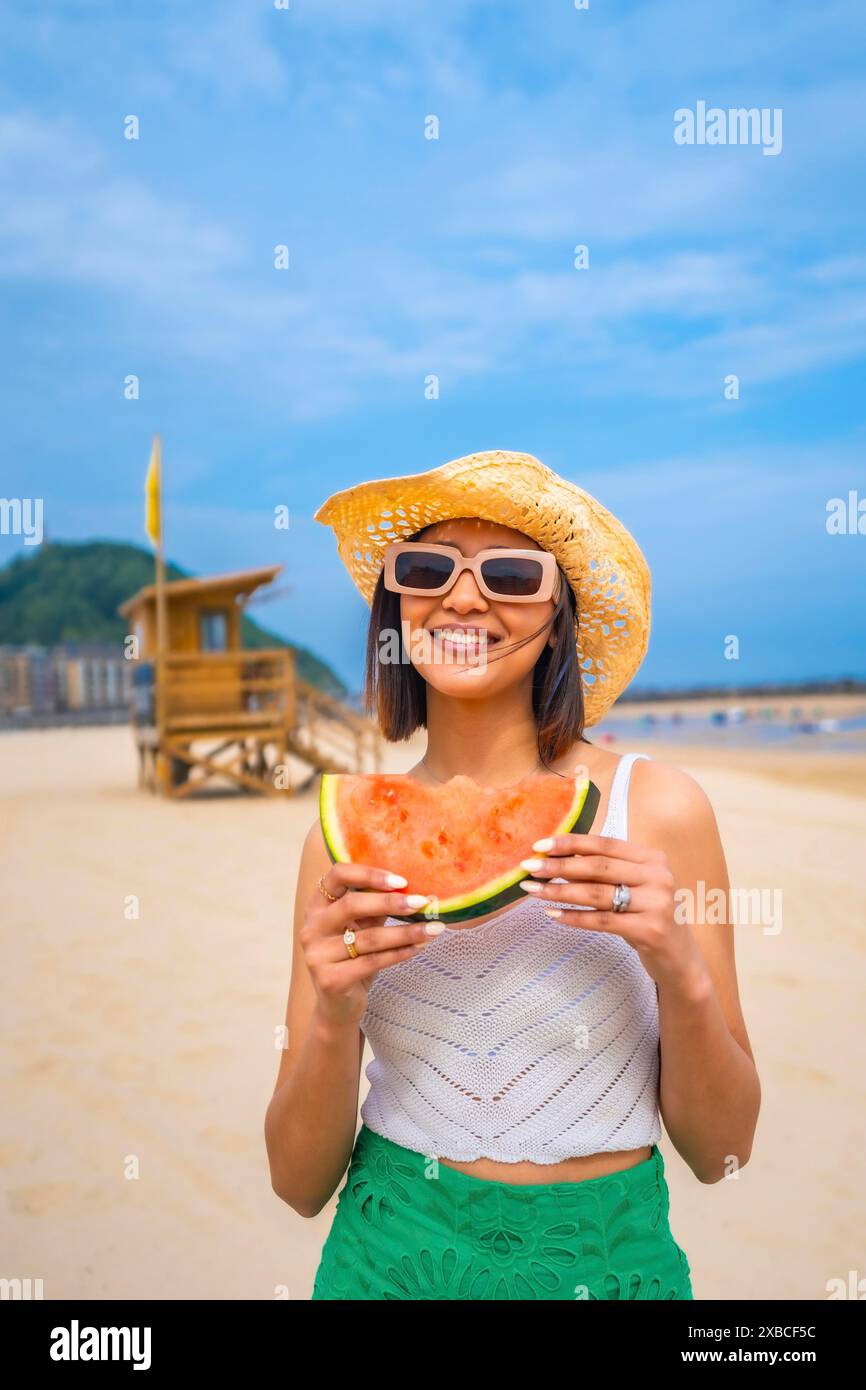 Una donna tiene in mano un anguria su una spiaggia. Lei sorride e indossa un cappello di paglia Foto Stock