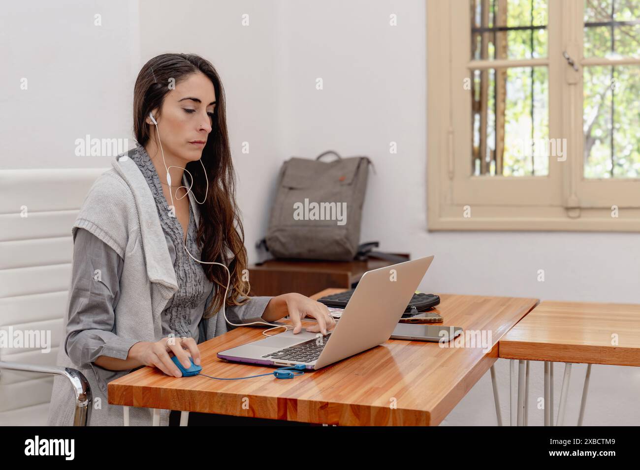 Una donna latina di mezza età, dai capelli lunghi, siede sulla scrivania dell'ufficio di casa, concentrandosi sul laptop mentre indossa le cuffie. Foto Stock