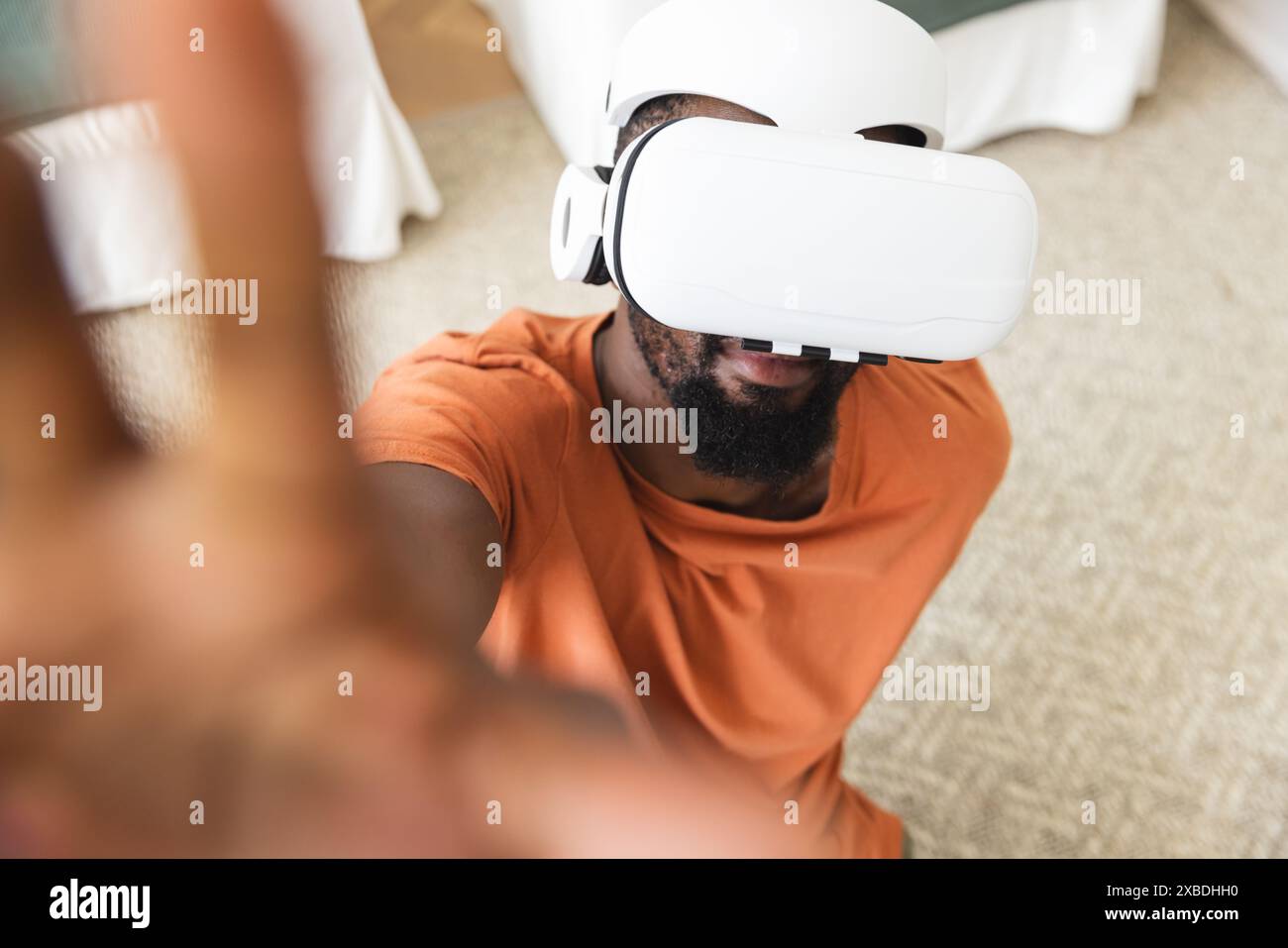 Giovane afroamericano che indossa un visore per la realtà virtuale, che si avvicina con la mano. E' in un soggiorno moderno con moquette beige e mobili bianchi, Foto Stock