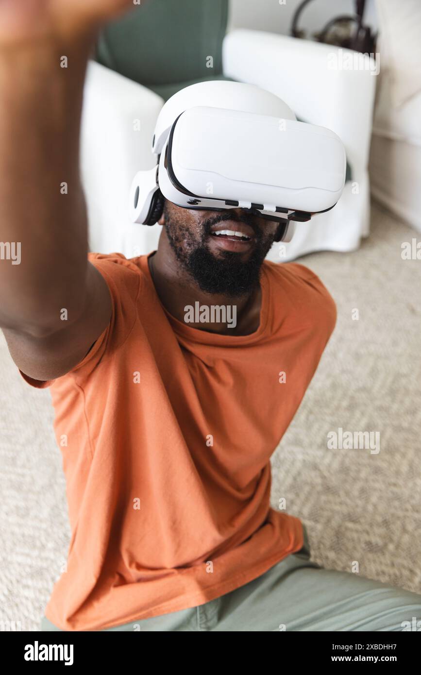 Giovane afroamericano con visore per la realtà virtuale che gli sta alzando la mano. Si trova in un accogliente soggiorno con un tappeto beige e mobili bianchi, un Foto Stock