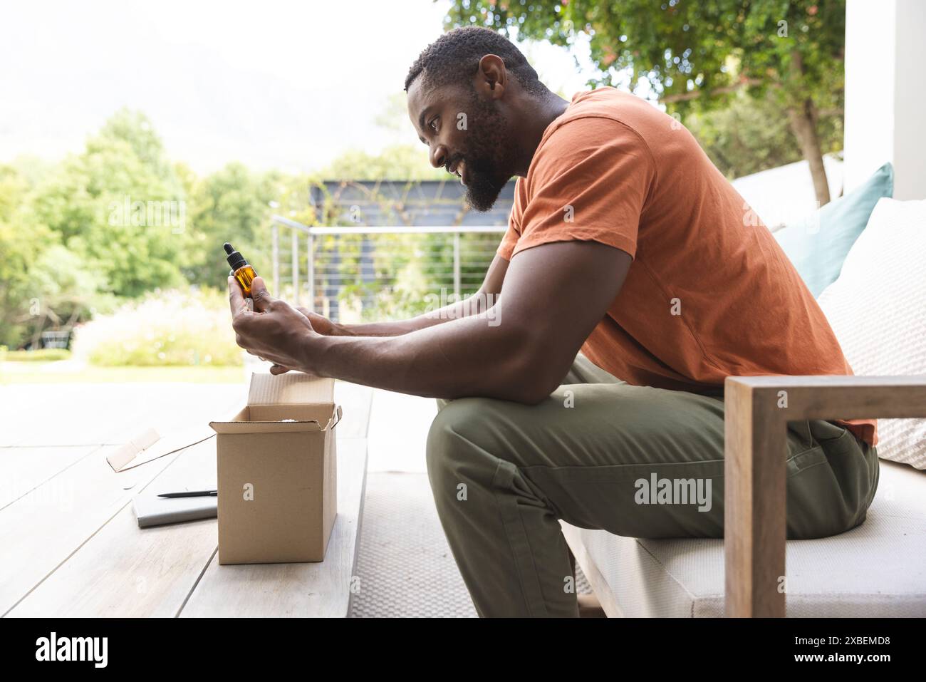 Giovane afroamericano sul patio pacchetto di apertura, esaminando una piccola bottiglia, a casa Foto Stock