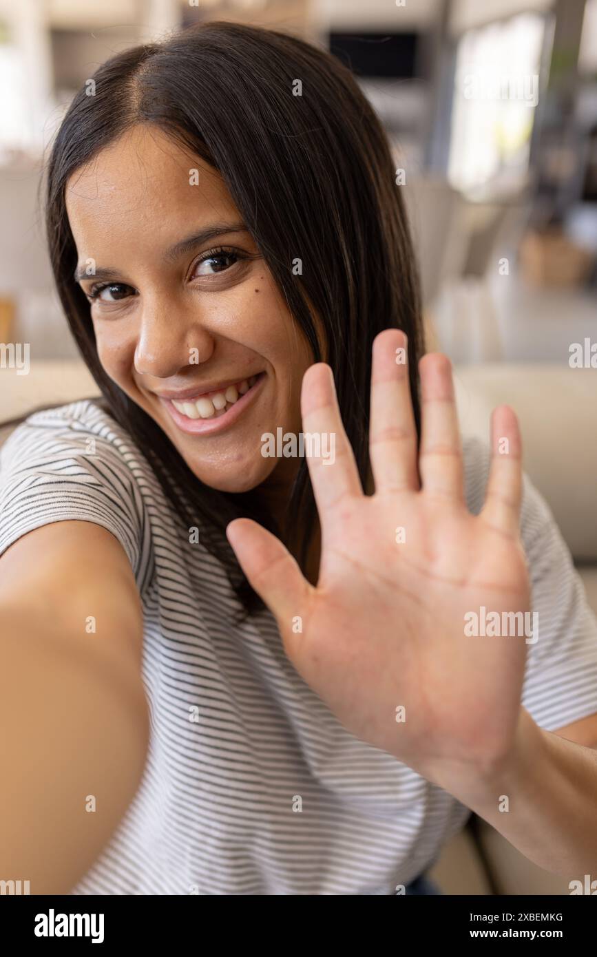 Giovane donna birazziale che sorride e sventola alla macchina fotografica, a casa Foto Stock