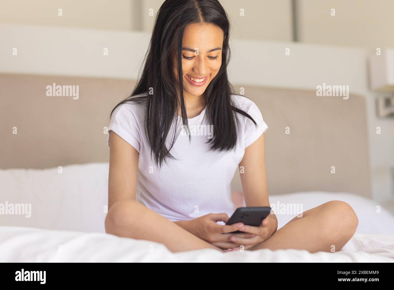 Giovane donna birazziale seduta sul letto, sorridente mentre guarda lo smartphone, a casa Foto Stock