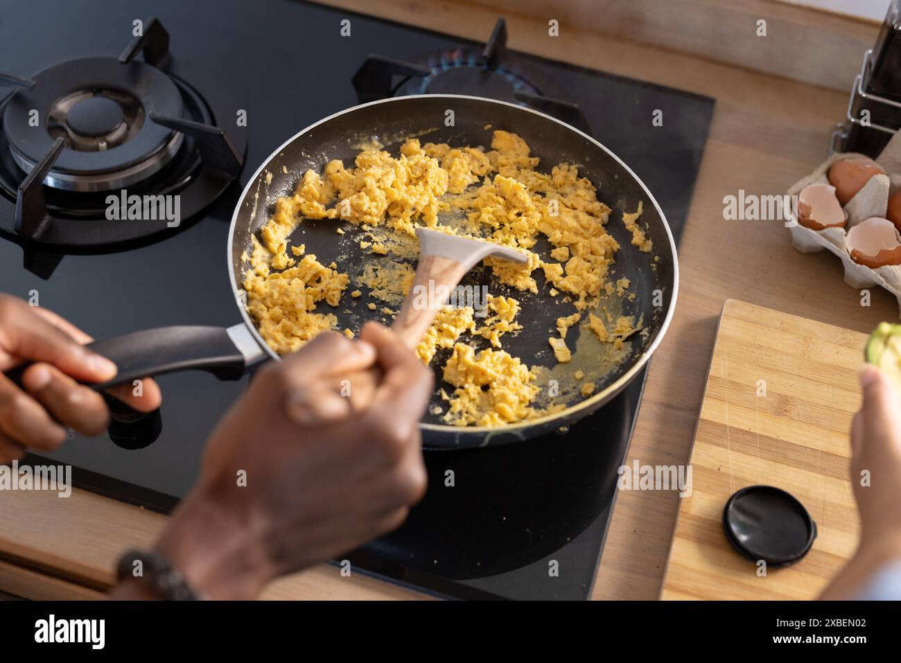 Una coppia variegata sta cucinando uova strapazzate insieme in una cucina moderna. Il piano di lavoro ha un tagliere con gusci d'uovo e avocado, creando un ambiente intimo Foto Stock