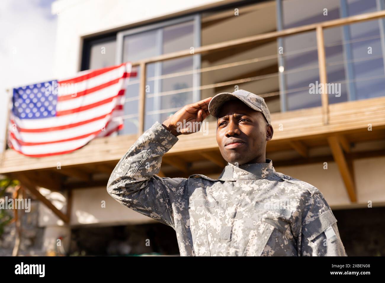 Giovane soldato afroamericano in uniforme militare che saluta. In piedi davanti alla casa con bandiera americana appesa sul balcone, mostrando orgoglio e sollievo Foto Stock