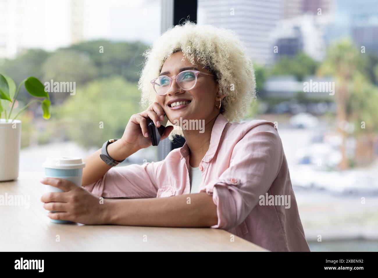 Una giovane donna birazziale con capelli ricci regala una tazza di caffè, parla sullo smartphone in ufficio Foto Stock