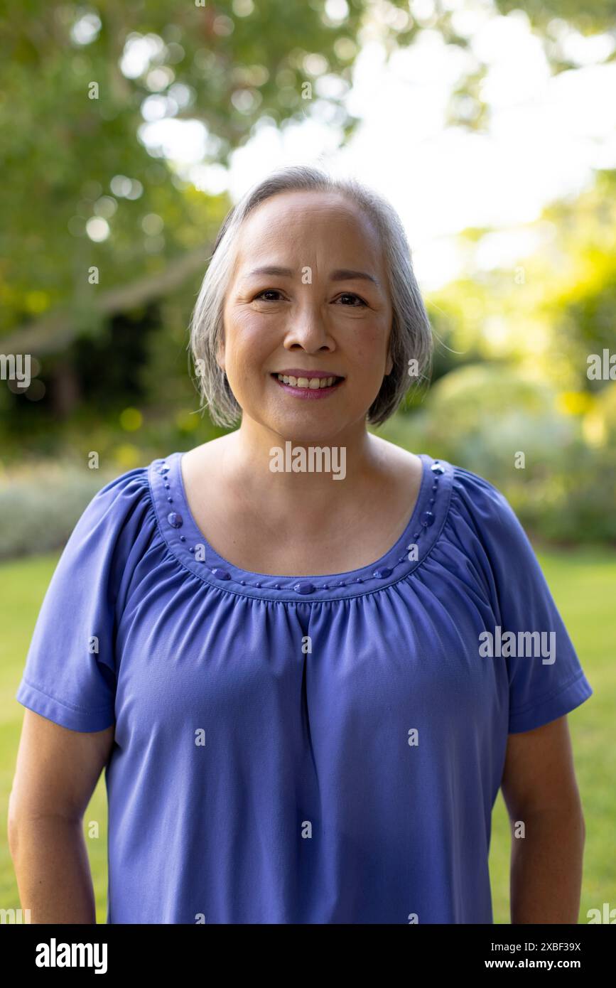 Donna asiatica senior sorridente calorosamente in giardino con vegetazione lussureggiante, all'aperto, a casa Foto Stock