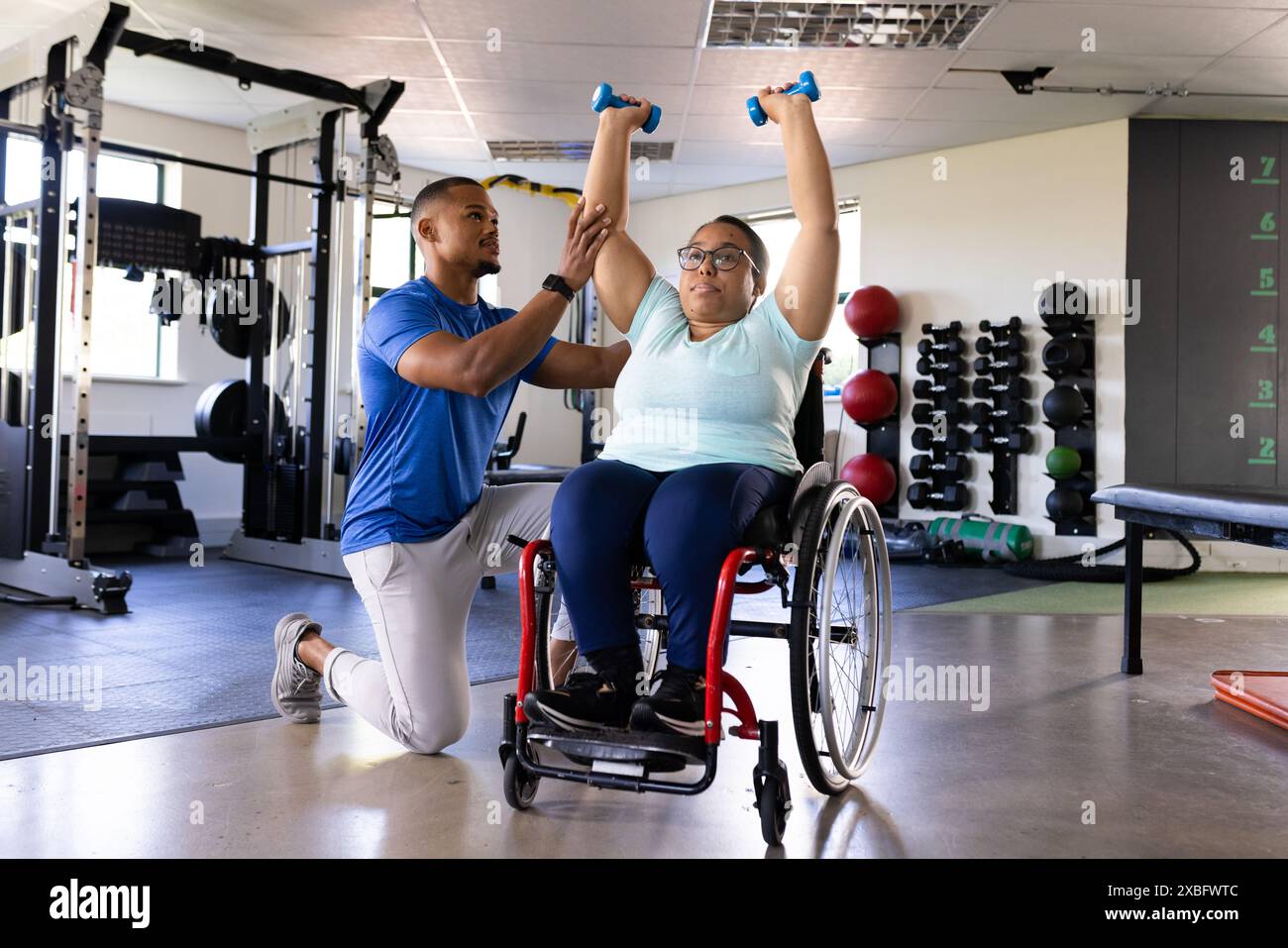 Donna quadriplegica birazziale in sedia a rotelle che si esercita con i manubri, assistita dall'allenatore sono in palestra riabilitativa con varie attrezzature per l'esercizio fisico Foto Stock