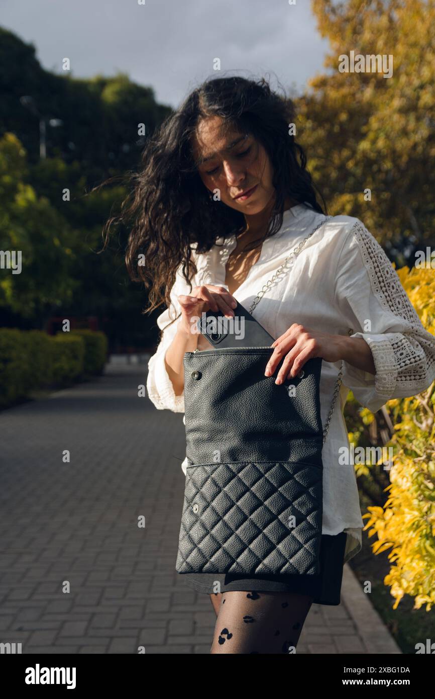 Una donna latina che prende il telefono dalla sua borsa sulla piazza. Indossa una camicia bianca e calze nere, immagine verticale, social media e techno Foto Stock