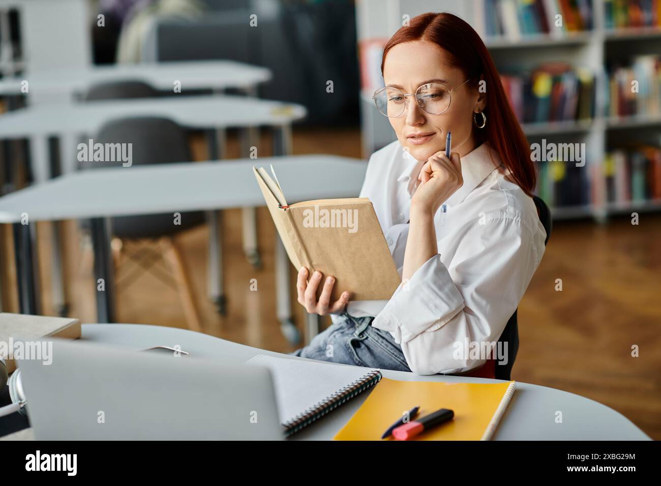 Una donna rossa immersa in un libro, circondata da scaffali di libri in una biblioteca, immersa in uno studio tranquillo. Foto Stock