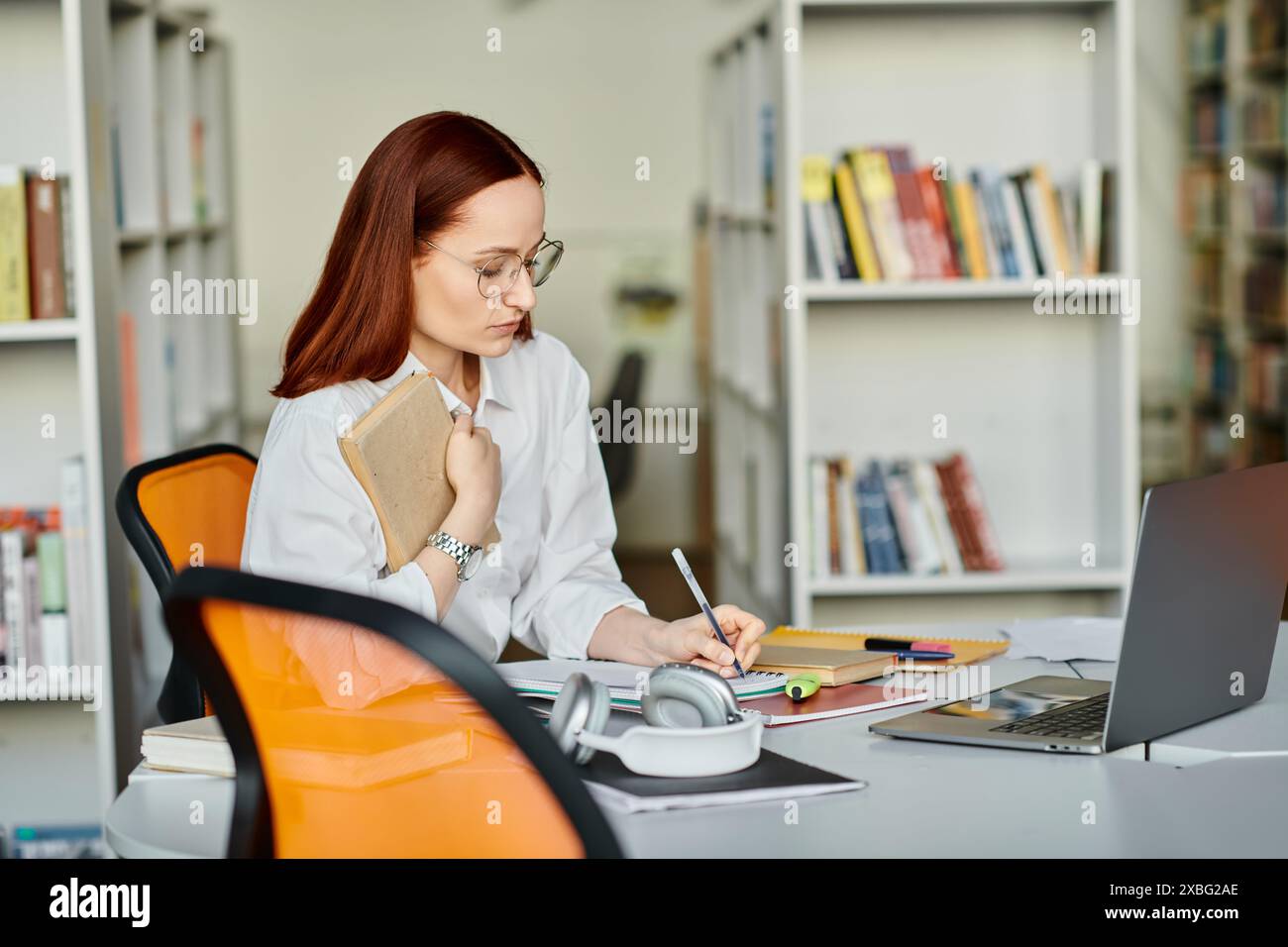 Una tutor donna con i capelli rossi che fornisce una lezione di after-School online, utilizzando un portatile a una scrivania in un ambiente libreria. Foto Stock