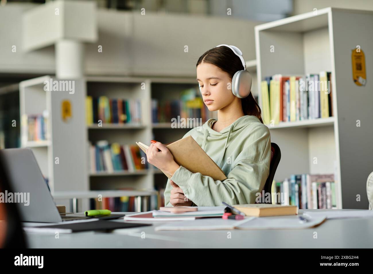 Una ragazza adolescente con le cuffie si siede a una scrivania in una biblioteca, immersa nel fare i compiti sul suo portatile dopo la scuola. Foto Stock