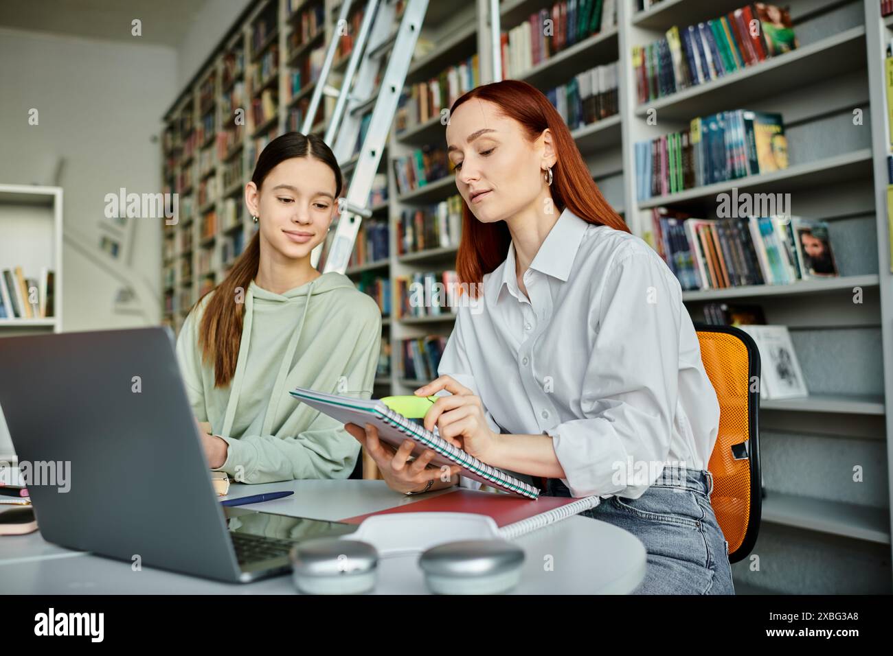 Sessione di tutoraggio in biblioteca, una tutor rossa e una ragazza adolescente lavorano su un notebook per l'istruzione moderna. Foto Stock