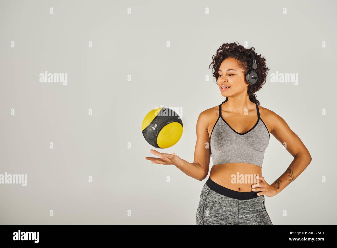 La sportiva riccia afroamericana in abbigliamento attivo mostra forza ed equilibrio mentre tiene in mano una palla gialla e nera vibrante. Foto Stock