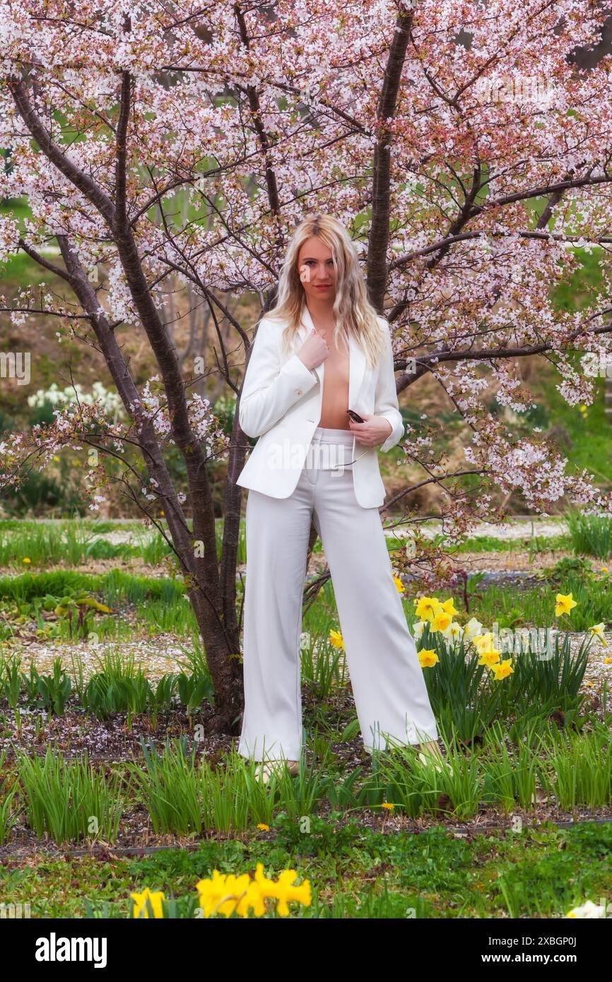 Una giovane donna bionda indossa elegantemente un pantalone bianco con una giacca aperta che mostra il suo petto. È circondata da un'abbondanza di fiori, Foto Stock