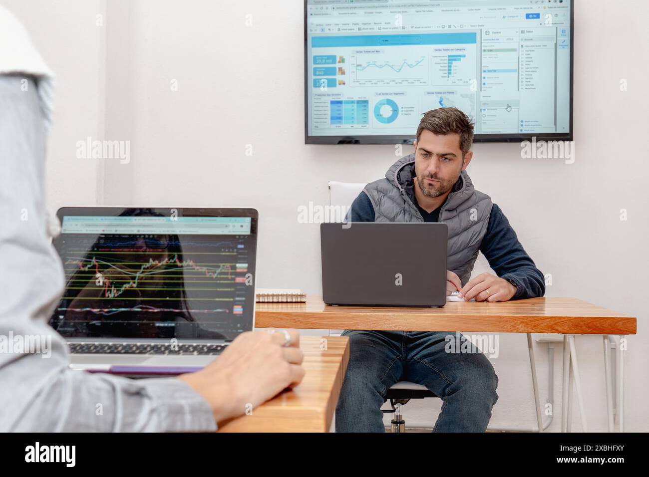 Un uomo di mezza età è profondamente concentrato sul suo notebook in un moderno spazio di coworking, con uno schermo che mostra grafica dietro di lui. Un'altra persona lavora vicino Foto Stock