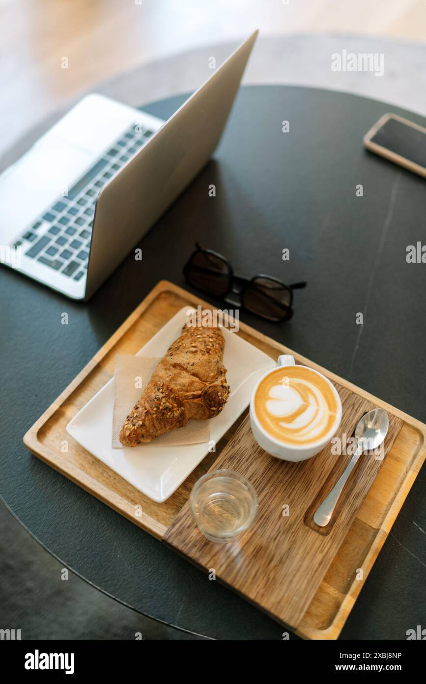 Un'immagine verticale di una scrivania da ufficio con un computer portatile, un caffè e un croissant su un vassoio Foto Stock