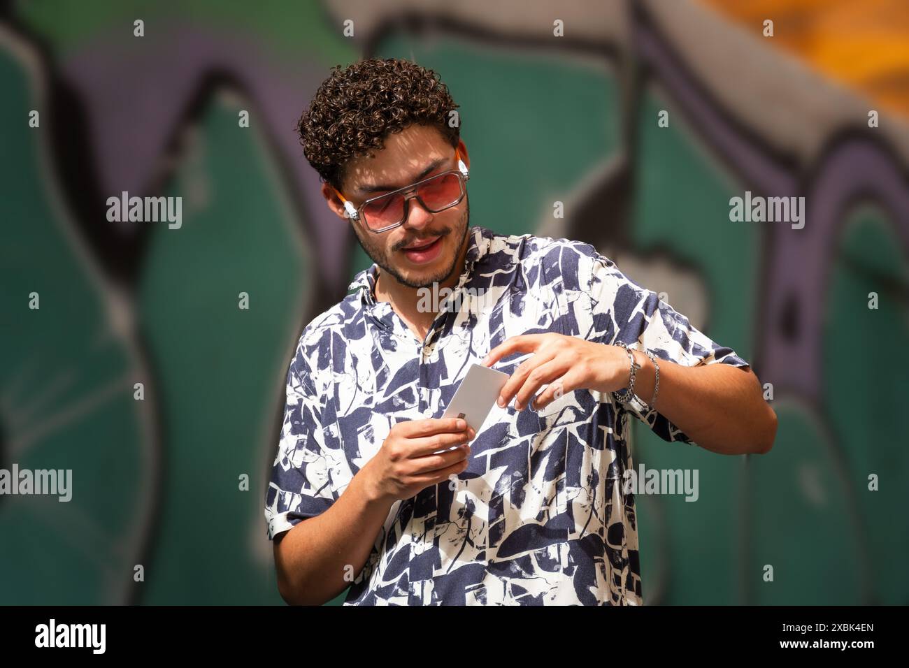 Giovane uomo moderno con occhiali da sole che guarda la sua carta di credito in città. Moda urbana, finanza, stile di vita Foto Stock