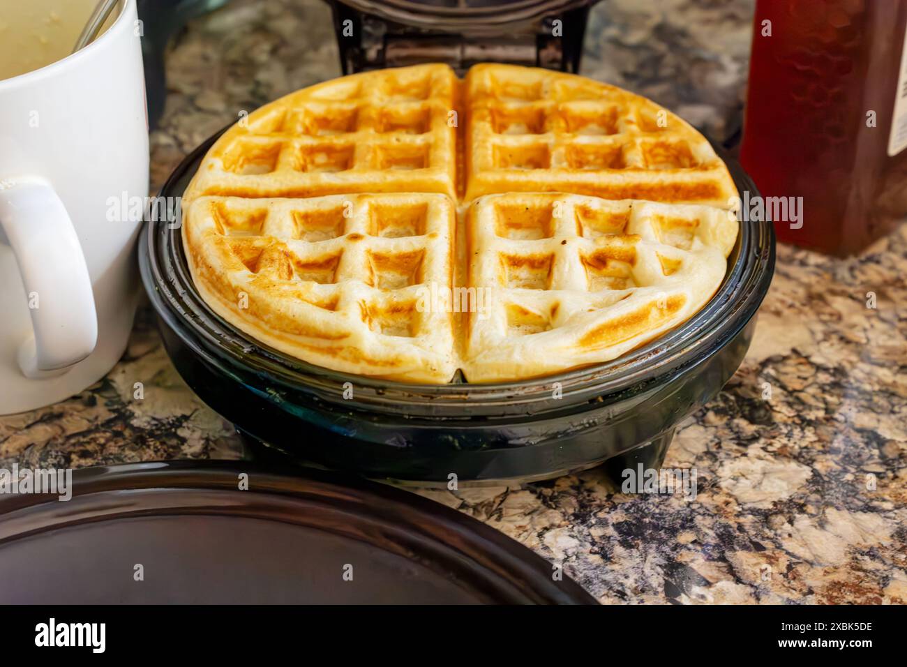 Sul ferro da stiro per cialde viene posizionato un waffle finito con una tazza per pastella e del miele sullo sfondo. Foto Stock