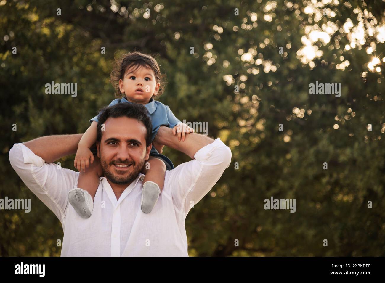 Un bambino di un anno seduto sulle spalle di suo padre con alberi e bokeh sullo sfondo, catturando un momento speciale nella festa del papà Foto Stock