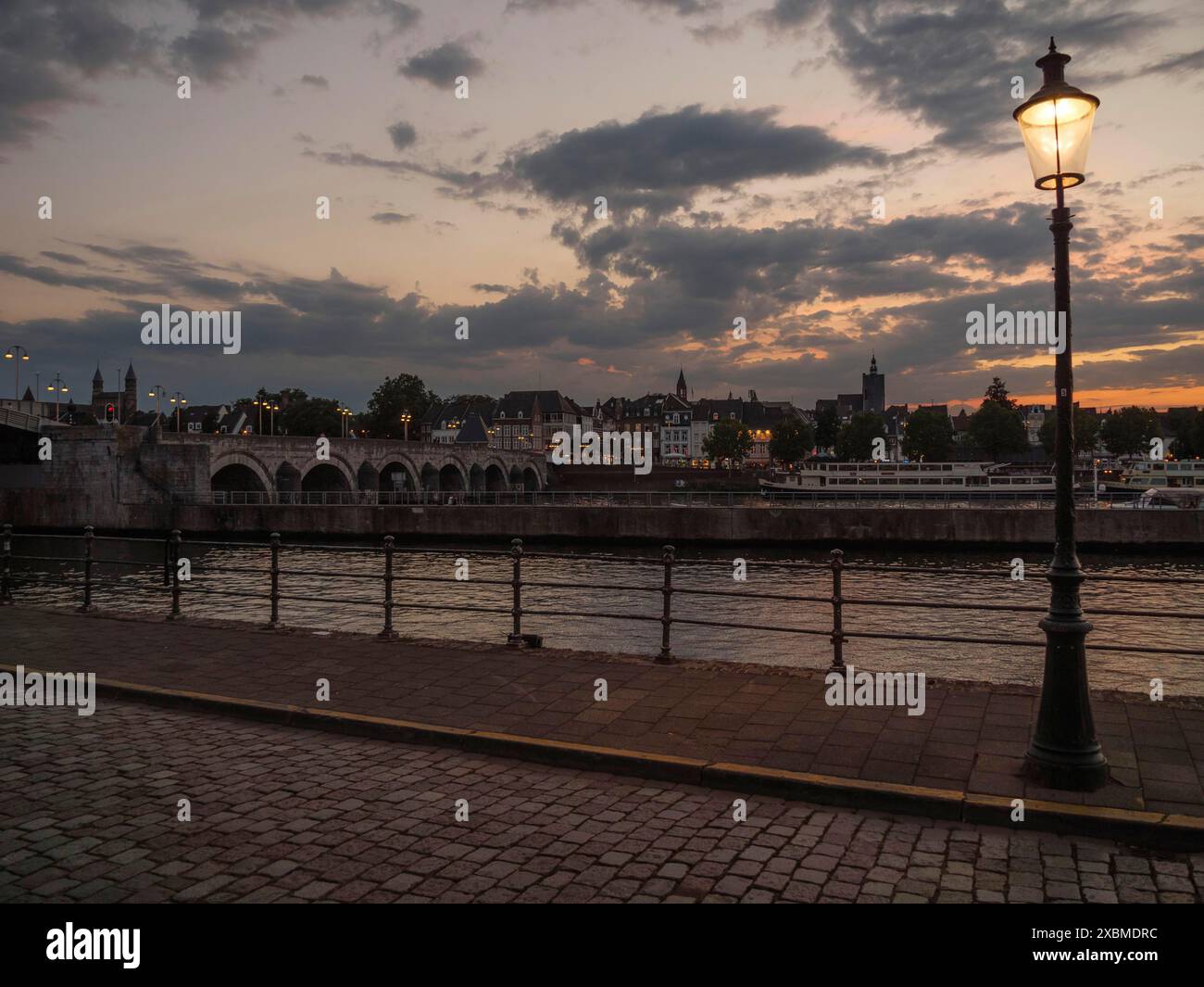 Riva del fiume al crepuscolo con un lampione stradale e una passerella acciottolata, Maastricht, limburgo, paesi bassi Foto Stock