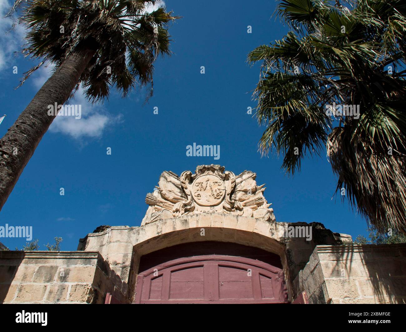 Struttura storica in pietra con uno stemma riccamente intagliato circondato da palme sotto un cielo azzurro, mdina, mediterraneo, malta Foto Stock
